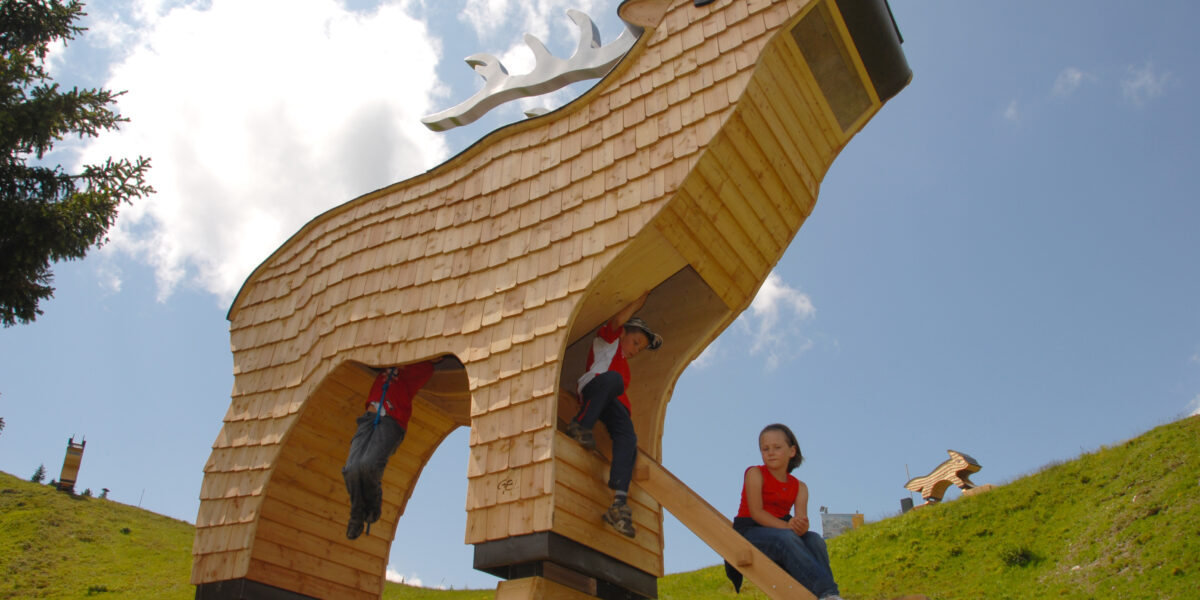 Große Holzkonstruktion in Form eines stilisierten Tieres, unter der Kinder klettern und spielen, auf einem grasbewachsenen Hügel unter einem teilweise bewölkten Himmel.