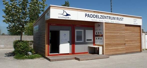 Ein kleines, modernes Holzgebäude mit einem Schild mit der Aufschrift „Paddelzentrum Rust“ und „seekajak“ an der Spitze, in dem sich ein Paddelsportzentrum befindet.