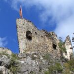 Die steinernen Ruinen einer alten Burg stehen auf einem felsigen Hügel, mit einer Fahne auf dem Gebäude und Bäumen, die das Gebiet unter einem teilweise bewölkten Himmel umgeben.