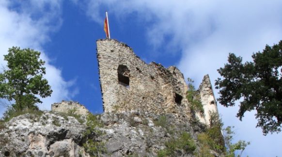 Die steinernen Ruinen einer alten Burg stehen auf einem felsigen Hügel, mit einer Fahne auf dem Gebäude und Bäumen, die das Gebiet unter einem teilweise bewölkten Himmel umgeben.