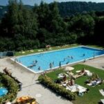 Das Freibad ist von Gras und Bäumen umgeben, und die Leute schwimmen im Pool, während andere sich sonnen oder auf den Liegestühlen in der Nähe sitzen.