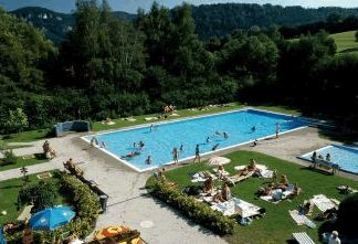 Das Freibad ist von Gras und Bäumen umgeben, und die Leute schwimmen im Pool, während andere sich sonnen oder auf den Liegestühlen in der Nähe sitzen.