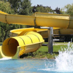 Eine große gelbe Wasserrutsche führt in ein Außenschwimmbecken, an dessen Ausgang das Wasser plätschert. Bäume und Gras umgeben den Poolbereich.