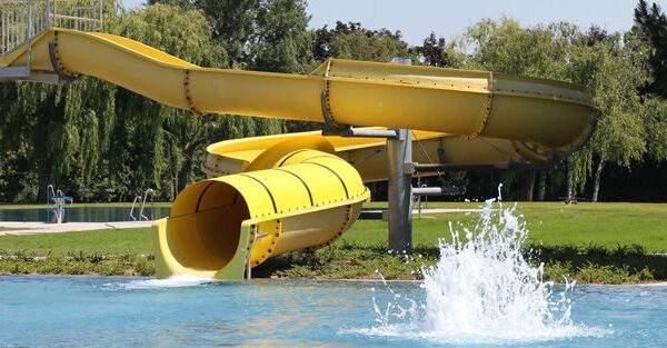 Eine große gelbe Wasserrutsche führt in ein Außenschwimmbecken, an dessen Ausgang das Wasser plätschert. Bäume und Gras umgeben den Poolbereich.