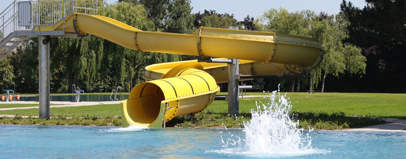 Eine große gelbe Wasserrutsche führt in ein Außenschwimmbecken, an dessen Ausgang das Wasser plätschert. Bäume und Gras umgeben den Poolbereich.