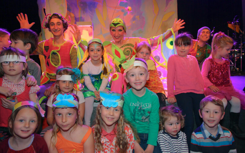 Kinder und zwei Erwachsene in bunten Kostümen sitzen und posieren für ein Gruppenfoto auf der Bühne, mit einer bunten Kulisse im Hintergrund.