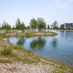 Ein kleiner See mit grasbewachsenen Ufern und vereinzelten Bäumen; an einem klaren Tag sitzen ein paar Leute am Wasser und entspannen sich.