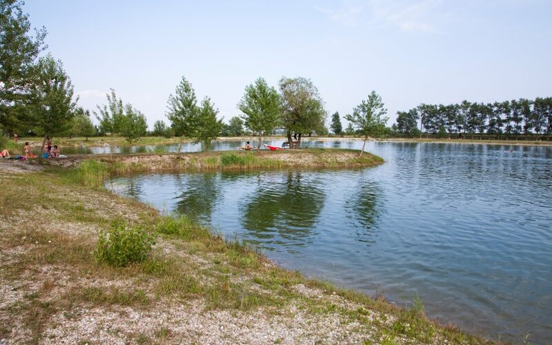 Ein kleiner See mit grasbewachsenen Ufern und vereinzelten Bäumen; an einem klaren Tag sitzen ein paar Leute am Wasser und entspannen sich.