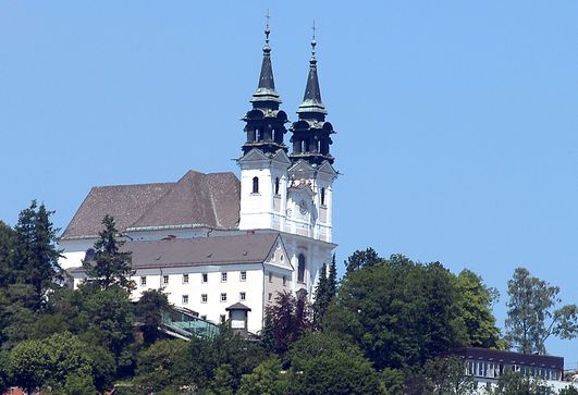 Eine weiße Kirche mit zwei hohen Türmen mit schwarzen Spitzen steht auf einem Hügel, umgeben von Bäumen unter einem klaren blauen Himmel.