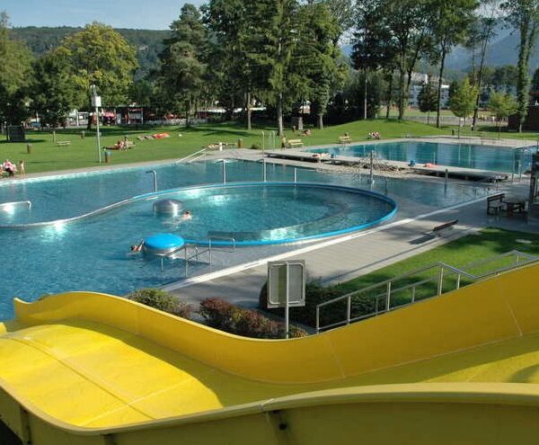 Freibadkomplex mit gelber Wasserrutsche, geschwungenen Beckenformen, Sonnenliegen auf der Wiese und umliegenden Bäumen und Hügeln.