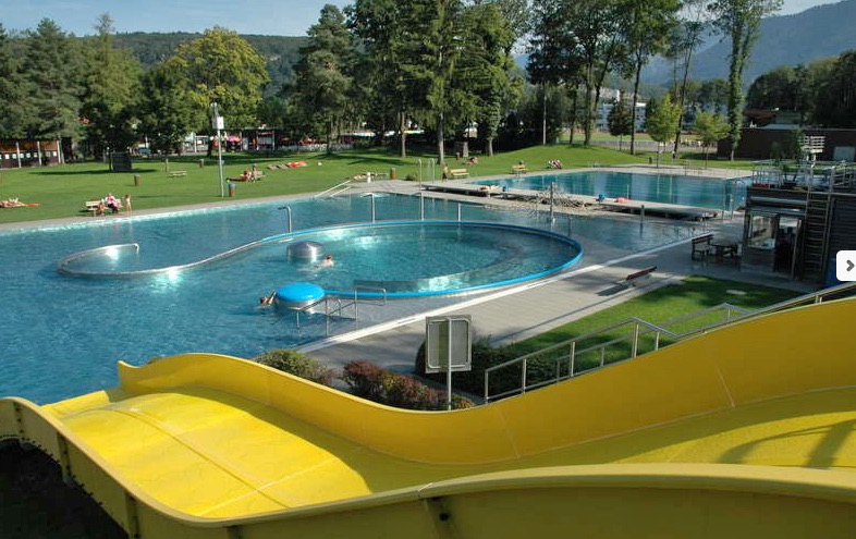 Freibadkomplex mit gelber Wasserrutsche, geschwungenen Beckenformen, Sonnenliegen auf der Wiese und umliegenden Bäumen und Hügeln.
