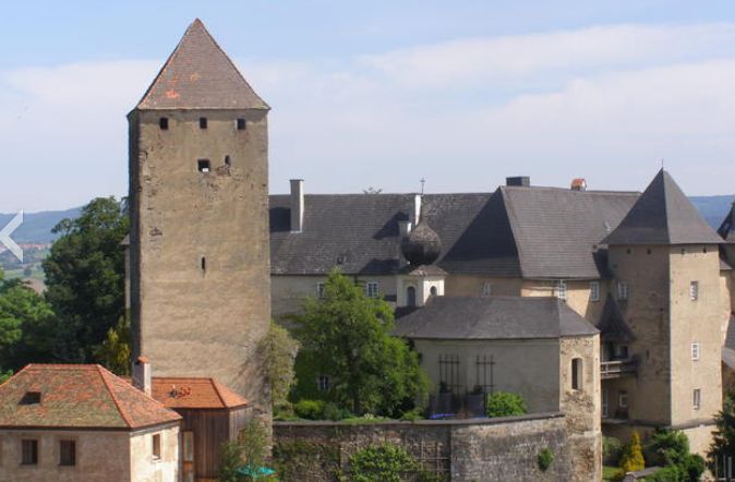 Steinburg mit einem hohen rechteckigen Turm, mehreren Satteldächern und umgebenden Bäumen vor einem blauen Himmel.