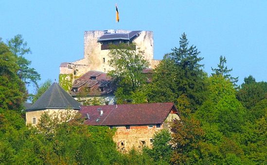 Auf einem Hügel steht eine Steinburg mit einer deutschen Flagge auf der Spitze, umgeben von Bäumen und mehreren kleineren Gebäuden mit roten Dachziegeln.