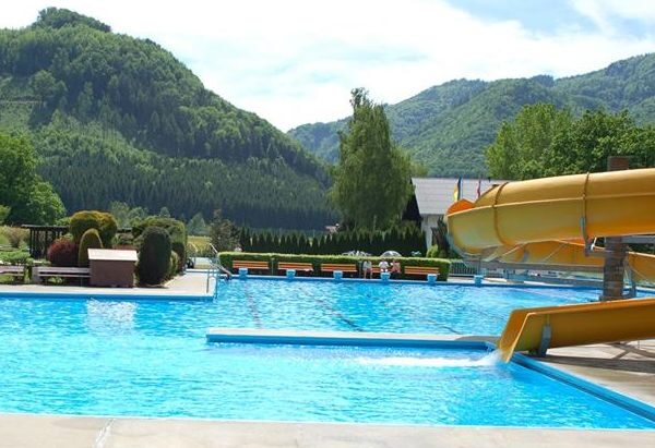 Außenpool mit gelber Wasserrutsche, umgeben von Grün und Bergen im Hintergrund unter einem teilweise bewölkten Himmel.