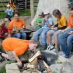 Eine Gruppe von Kindern und ein Erwachsener sitzen im Freien um eine Feuerstelle, einige halten Stöcke in der Hand, während ein Junge auf das unangezündete Holz pustet, um ein Lagerfeuer zu entfachen.