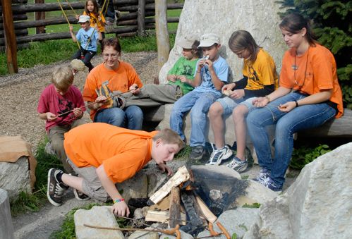 Eine Gruppe von Kindern und ein Erwachsener sitzen im Freien um eine Feuerstelle, einige halten Stöcke in der Hand, während ein Junge auf das unangezündete Holz pustet, um ein Lagerfeuer zu entfachen.