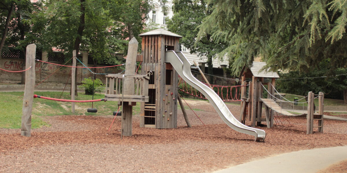 Ein hölzernes Spielgerät mit Rutsche, Seilbrücken und Kletterelementen, das auf Mulch in einem Park mit Bäumen und einem gepflasterten Weg in der Nähe steht.