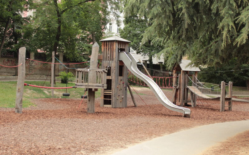 Ein hölzernes Spielgerät mit Rutsche, Seilbrücken und Kletterelementen, das auf Mulch in einem Park mit Bäumen und einem gepflasterten Weg in der Nähe steht.