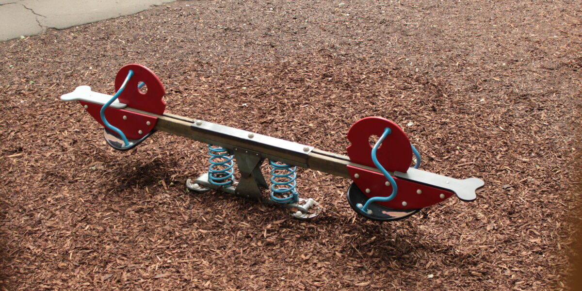 Eine Wippe mit roten Sitzen und blauen Federn steht auf einem mit Holzspänen bedeckten Spielplatz.