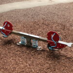 Eine Wippe mit roten Sitzen und blauen Federn steht auf einem mit Holzspänen bedeckten Spielplatz.