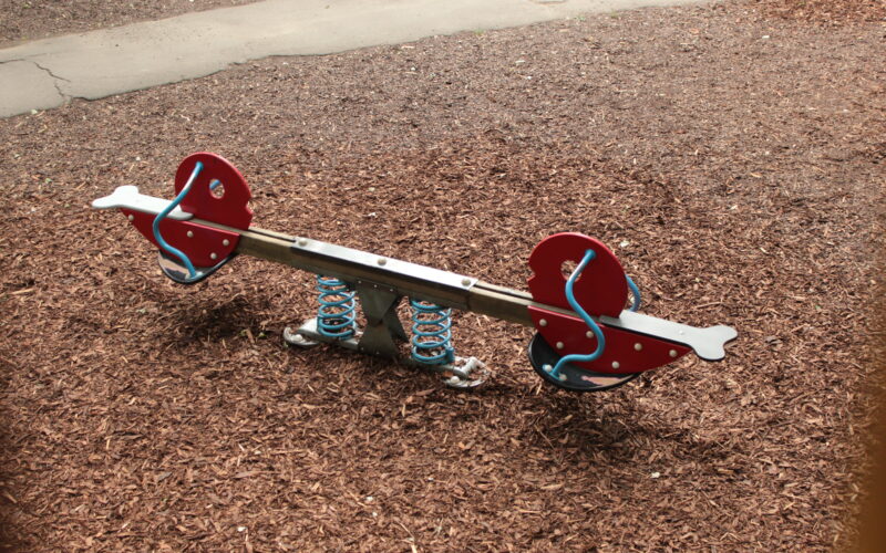 Eine Wippe mit roten Sitzen und blauen Federn steht auf einem mit Holzspänen bedeckten Spielplatz.