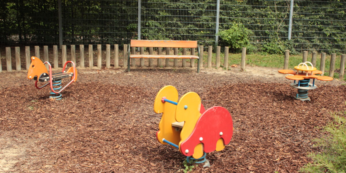 Ein Spielplatz mit tierförmigen Springreitern, einem Drehsitz, einer Holzbank und einem Zaun im Hintergrund.