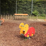 Ein Spielplatz mit tierförmigen Springreitern, einem Drehsitz, einer Holzbank und einem Zaun im Hintergrund.