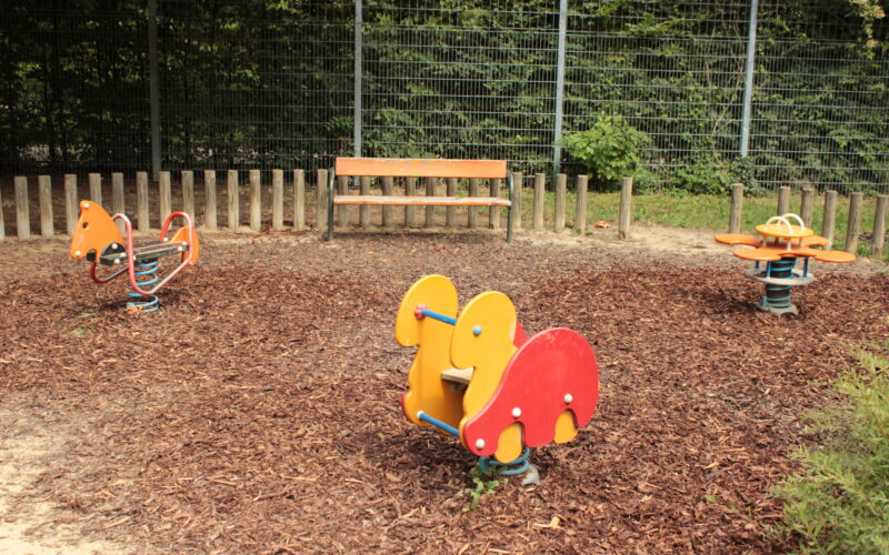 Ein Spielplatz mit tierförmigen Springreitern, einem Drehsitz, einer Holzbank und einem Zaun im Hintergrund.