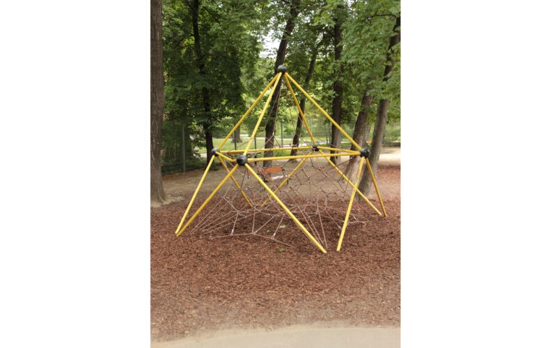 Ein gelbes Metallklettergerüst mit Seilnetz steht auf Raufaserboden in einem von Bäumen umgebenen Park.