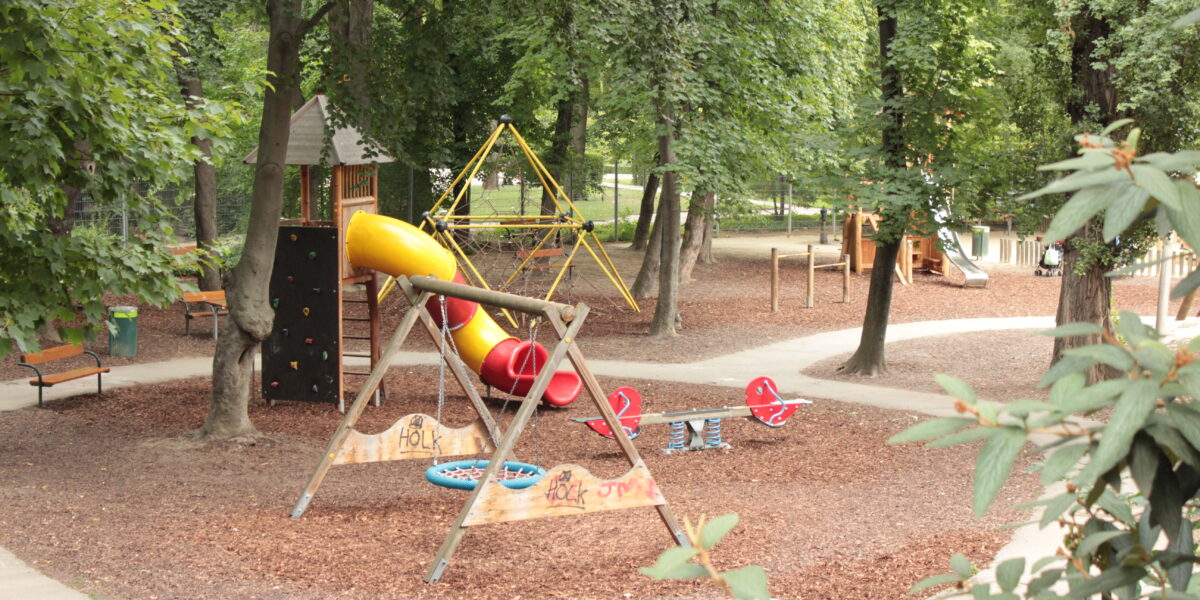 Spielplatz in einem Park mit Rutsche, Kletterwand, Schaukeln und Springreitern, umgeben von Bäumen und Wegen.