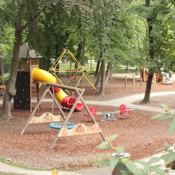 Spielplatz in einem Park mit Rutsche, Kletterwand, Schaukeln und Springreitern, umgeben von Bäumen und Wegen.