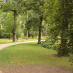 An einem bewölkten Tag führt ein geschwungener Schotterweg durch einen grünen Park mit hohen Bäumen, Gras und Büschen.