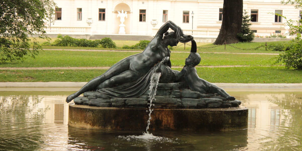 Eine bronzene Brunnenskulptur mit zwei liegenden Figuren, aus deren Hand Wasser fließt, in einem Teich vor einem großen weißen Gebäude.