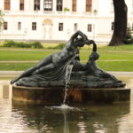 Eine bronzene Brunnenskulptur mit zwei liegenden Figuren, aus deren Hand Wasser fließt, in einem Teich vor einem großen weißen Gebäude.