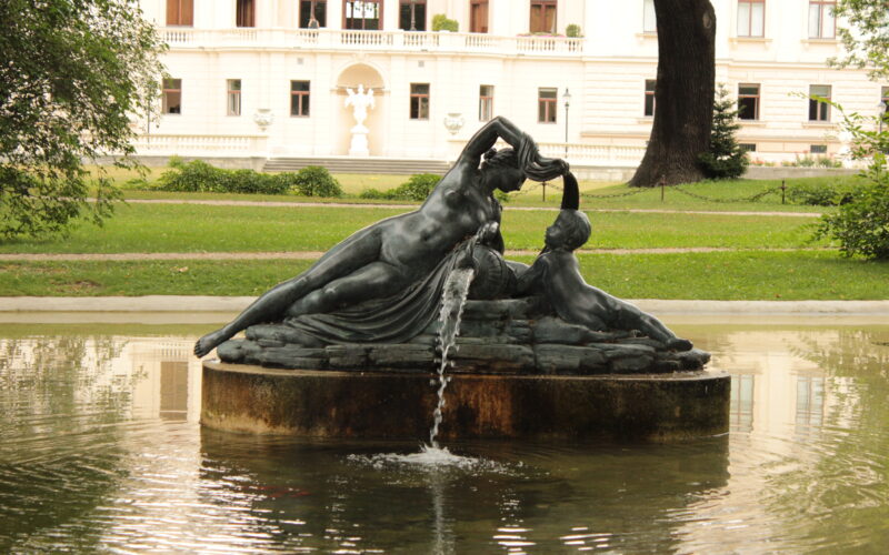 Eine bronzene Brunnenskulptur mit zwei liegenden Figuren, aus deren Hand Wasser fließt, in einem Teich vor einem großen weißen Gebäude.