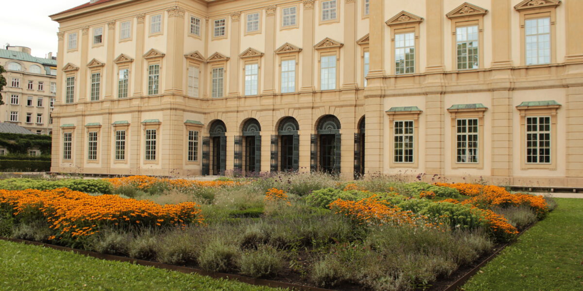Hinter einem angelegten Garten mit orangefarbenen und violetten Blumen und grünem Gras steht ein großes beigefarbenes historisches Gebäude mit vielen Fenstern.