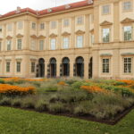 Hinter einem angelegten Garten mit orangefarbenen und violetten Blumen und grünem Gras steht ein großes beigefarbenes historisches Gebäude mit vielen Fenstern.