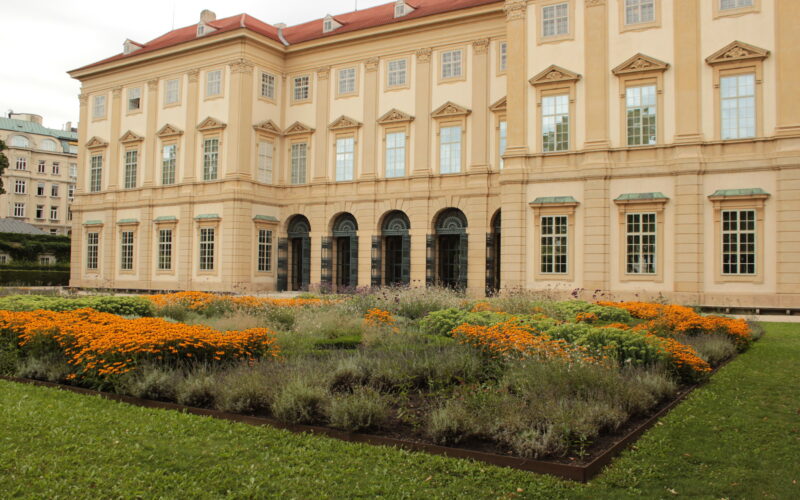 Hinter einem angelegten Garten mit orangefarbenen und violetten Blumen und grünem Gras steht ein großes beigefarbenes historisches Gebäude mit vielen Fenstern.