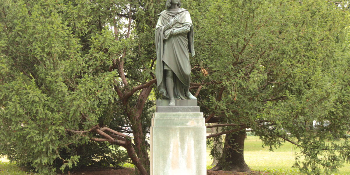 Eine Bronzestatue einer gekleideten Figur steht auf einem rechteckigen Sockel vor grünen Bäumen im Freien.