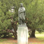 Eine Bronzestatue einer gekleideten Figur steht auf einem rechteckigen Sockel vor grünen Bäumen im Freien.