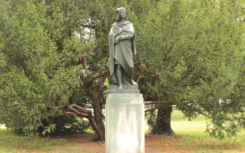 Eine Bronzestatue einer gekleideten Figur steht auf einem rechteckigen Sockel vor grünen Bäumen im Freien.