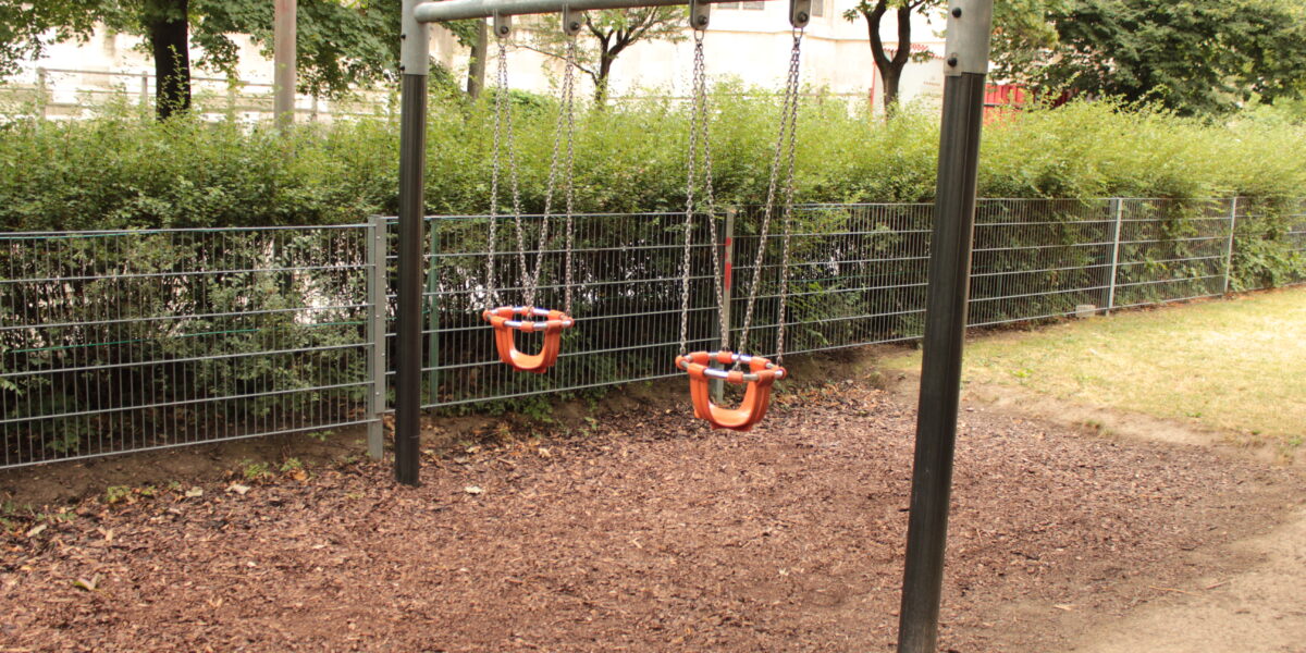 Zwei leere Kleinkinderschaukeln mit Sicherheitssitzen hängen an einem Metallrahmen in einem Spielplatz im Freien mit Raufaserbelag.