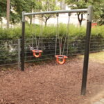 Zwei leere Kleinkinderschaukeln mit Sicherheitssitzen hängen an einem Metallrahmen in einem Spielplatz im Freien mit Raufaserbelag.