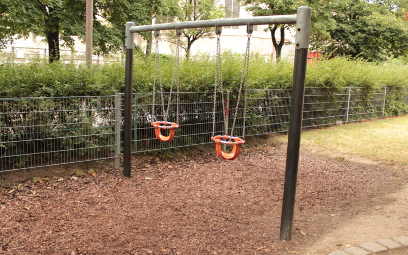 Zwei leere Kleinkinderschaukeln mit Sicherheitssitzen hängen an einem Metallrahmen in einem Spielplatz im Freien mit Raufaserbelag.