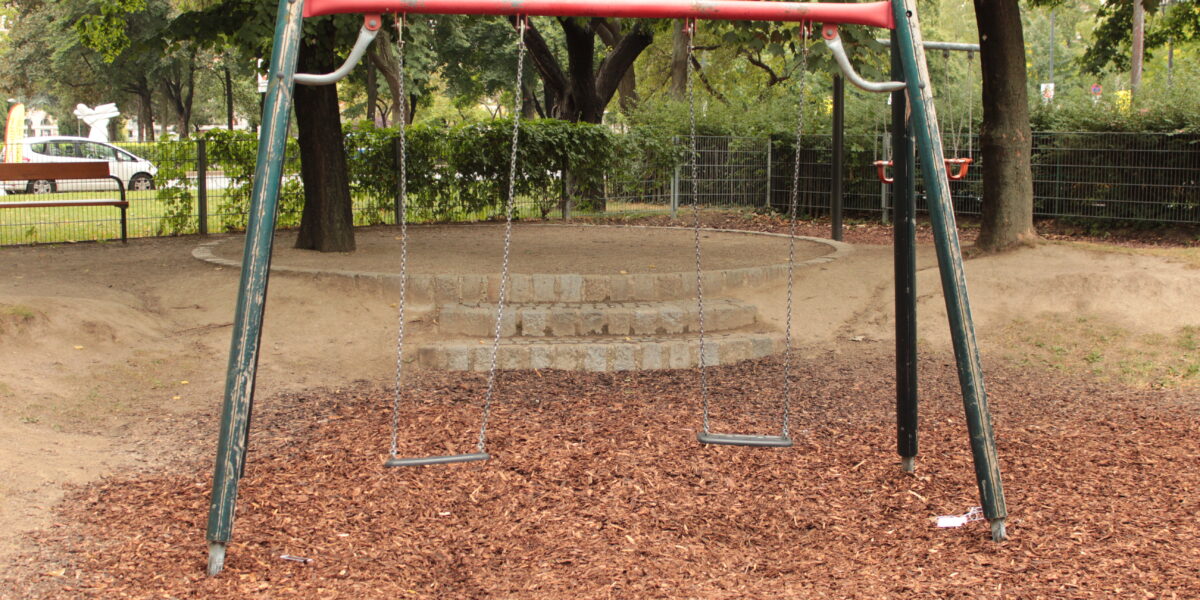 Eine leere Schaukel mit zwei Sitzen steht auf Holzspänen in einem Park, umgeben von Bäumen und einem eingezäunten Gelände im Hintergrund.