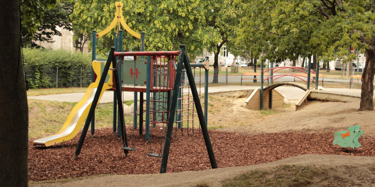 Ein Spielplatz mit einer gelben Rutsche, einem Klettergerüst und einer Schaukel liegt auf Mulch, neben einer kleinen Holzbrücke und einer grünen Federwippe, umgeben von Bäumen.