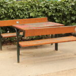 Holzbänke und ein Tisch mit Metallbeinen stehen auf einer Betonfläche im Freien, im Hintergrund eine grüne, belaubte Hecke.