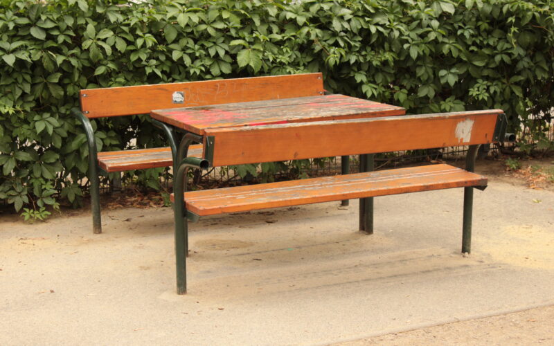 Holzbänke und ein Tisch mit Metallbeinen stehen auf einer Betonfläche im Freien, im Hintergrund eine grüne, belaubte Hecke.