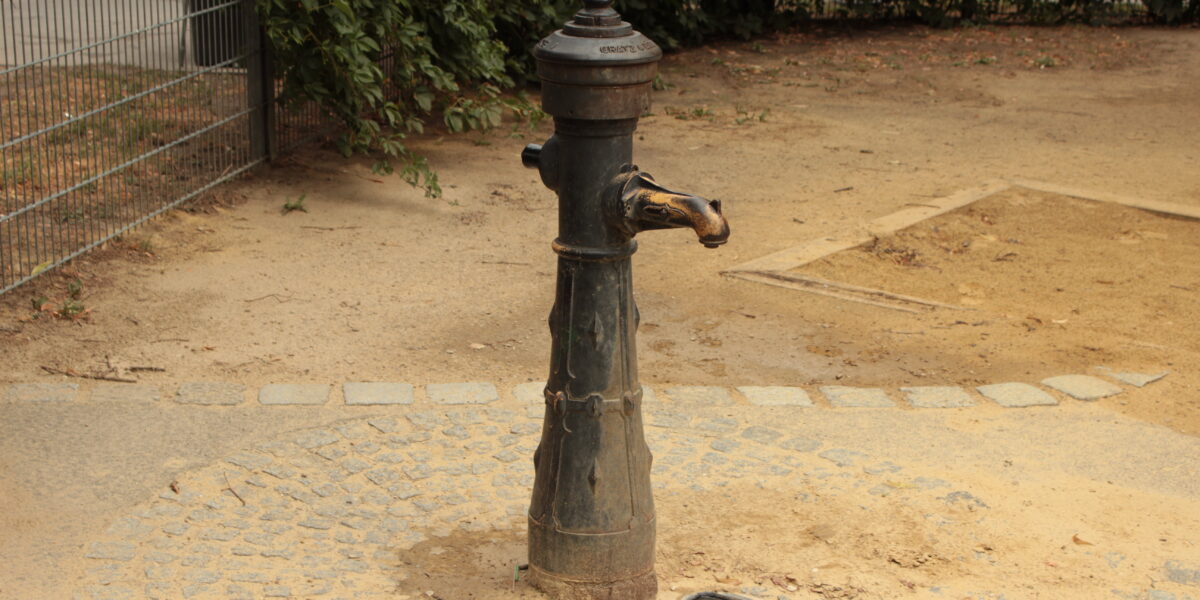 Eine alte Wasserpumpe steht auf einer runden Steinfläche, umgeben von Erde, mit Büschen und einem Metallzaun im Hintergrund.