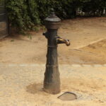 Eine alte Wasserpumpe steht auf einer runden Steinfläche, umgeben von Erde, mit Büschen und einem Metallzaun im Hintergrund.